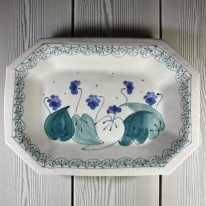 May include: White ceramic serving platter with a hand-painted floral design. The rectangular dish features a border of teal scrollwork and a central motif of blue flowers and green leaves. The platter is approximately 12 inches long and 8 inches wide.