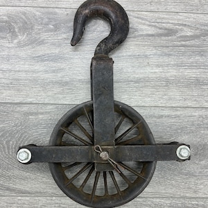 May include: An antique, cast iron pulley with a large hook and a spoked wheel. The metal has a weathered, dark gray finish, showing signs of age and use. The pulley is designed for heavy-duty lifting and is mounted on a wooden surface.