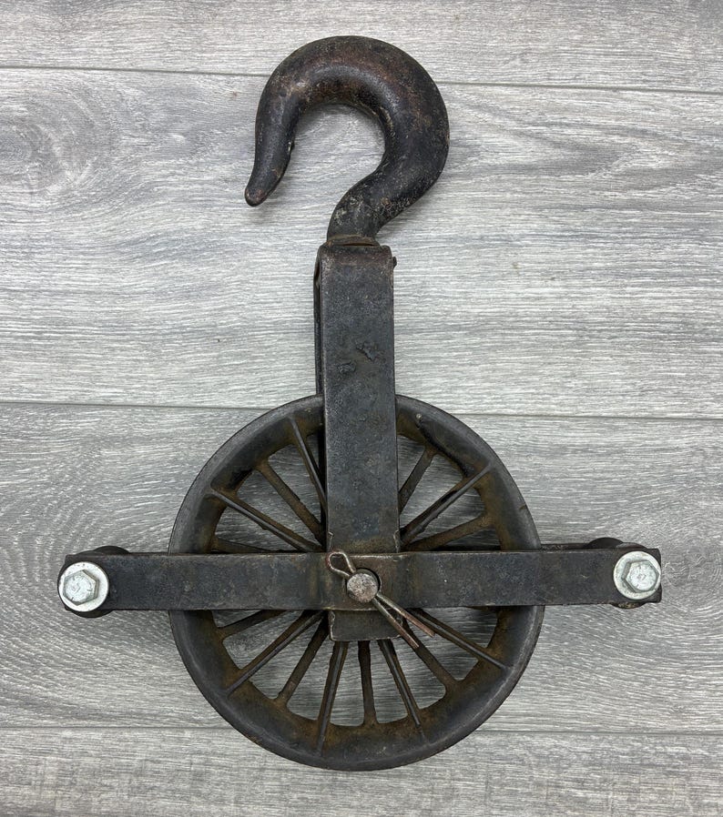 May include: An antique, cast iron pulley with a large hook and a spoked wheel. The metal has a weathered, dark gray finish, showing signs of age and use. The pulley is designed for heavy-duty lifting and is mounted on a wooden surface.