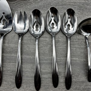 May include: A set of six stainless steel serving utensils, including a cake server, fork, slotted spoons, and a ladle. Each piece has a decorative cut-out design and a dark, patterned handle. The utensils are arranged on a gray wooden surface.