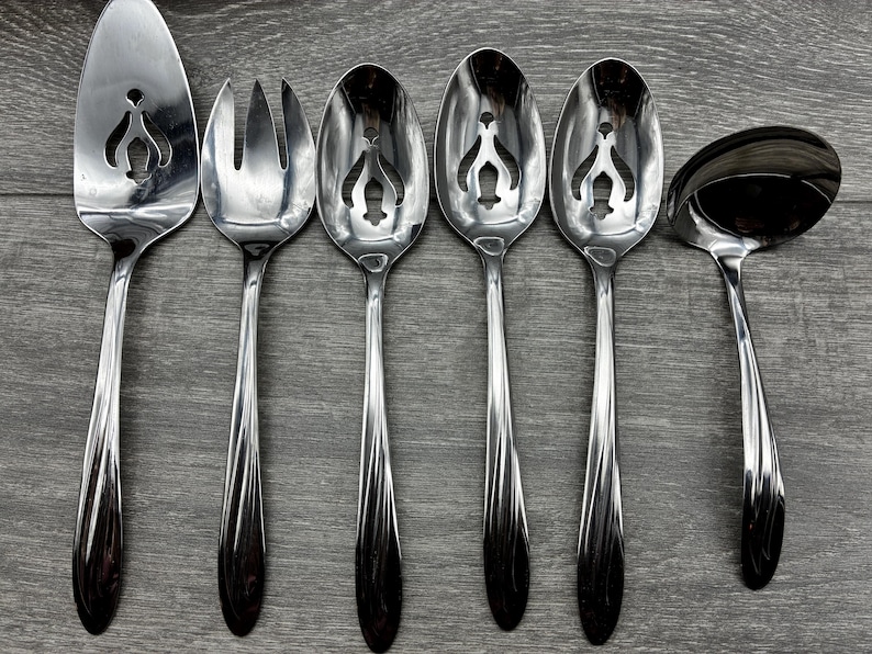 May include: A set of six stainless steel serving utensils, including a cake server, fork, slotted spoons, and a ladle. Each piece has a decorative cut-out design and a dark, patterned handle. The utensils are arranged on a gray wooden surface.