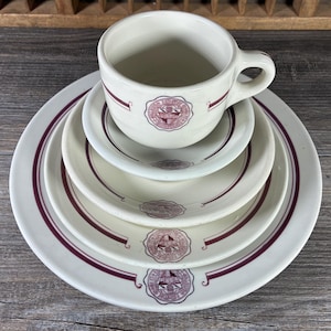 May include: A cream-colored vintage dish set with a cup, saucer, and three plates. Each piece features a burgundy border and a central emblem. The cup is stacked on the saucer and plates, creating a layered effect. The set is arranged on a wooden surface.