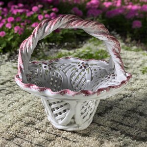 May include: White ceramic basket with a pink and white twisted handle. The basket has a woven design and floral accents.