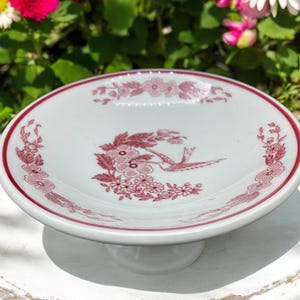 May include: A small white ceramic pedestal cake stand with a red floral design and a bird in the center.