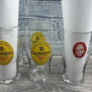 K&ouml;nnte beinhalten: Mehrere klare Guinness-Biergl&auml;ser mit Papiereinlagen. Die Gl&auml;ser haben gelbe und schwarze Guinness-Etiketten. Ein Glas hat ein rot-wei&szlig;es Guinness Porter-Etikett. Die Gl&auml;ser stehen auf einer grauen Holzoberfl&auml;che.