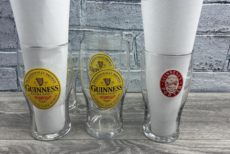 K&ouml;nnte beinhalten: Mehrere klare Guinness-Biergl&auml;ser mit Papiereinlagen. Die Gl&auml;ser haben gelbe und schwarze Guinness-Etiketten. Ein Glas hat ein rot-wei&szlig;es Guinness Porter-Etikett. Die Gl&auml;ser stehen auf einer grauen Holzoberfl&auml;che.