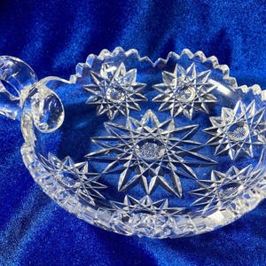 May include: A clear glass dish with a starburst pattern and a handle. The dish is perfect for serving snacks or desserts.