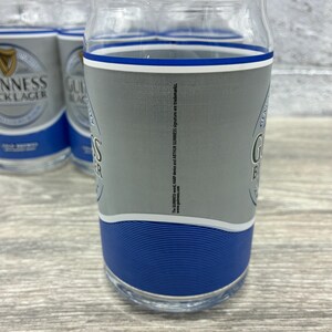 May include: Clear glass pint glasses with a gray and blue design. The glasses feature the Guinness logo and branding. The glasses are cylindrical with a slightly curved top and a blue band at the base.