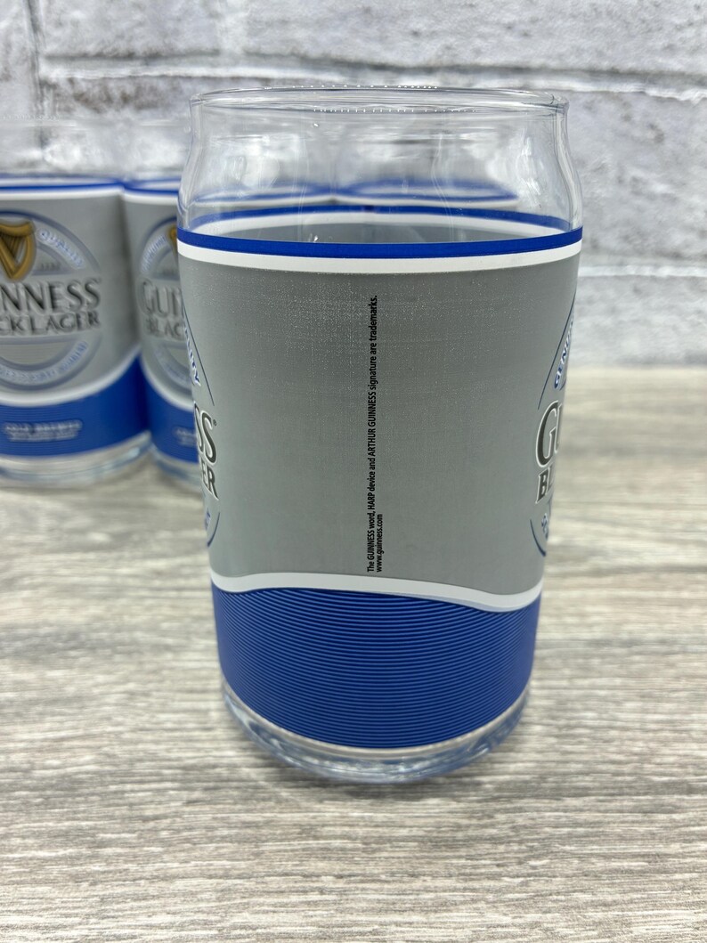 May include: Clear glass pint glasses with a gray and blue design. The glasses feature the Guinness logo and branding. The glasses are cylindrical with a slightly curved top and a blue band at the base.