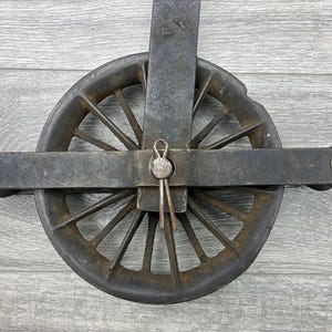 May include: An antique, cast iron pulley with a wheel featuring multiple spokes. The pulley has a dark, weathered appearance with a central pin and two bolts on either side. The wheel is approximately 6 inches in diameter.