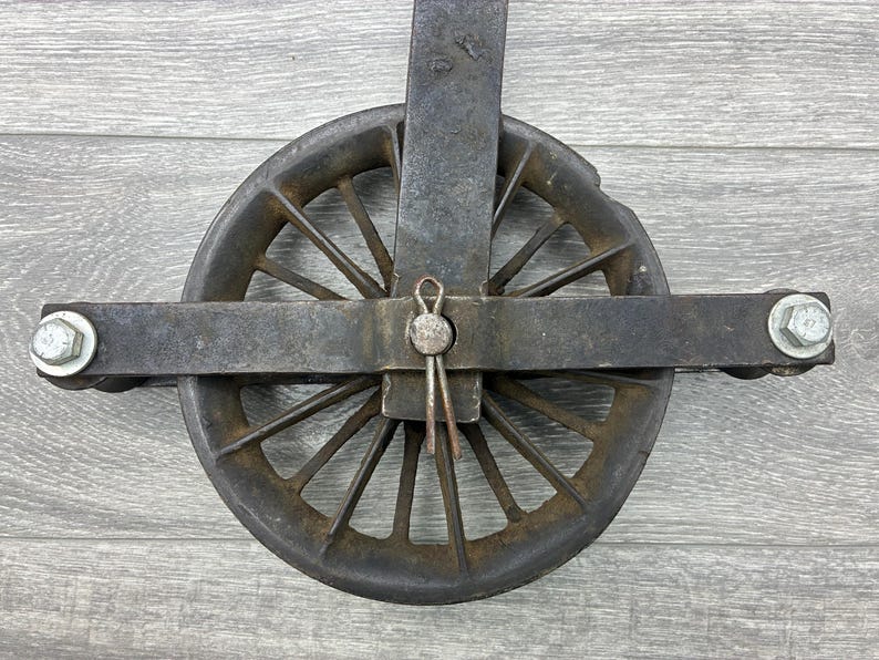 May include: An antique, cast iron pulley with a wheel featuring multiple spokes. The pulley has a dark, weathered appearance with a central pin and two bolts on either side. The wheel is approximately 6 inches in diameter.