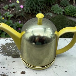 May include: A teapot featuring a polished gold-tone metal body with a bright yellow handle, spout, and lid knob. The lid is domed and metallic, while the spout is angled and the handle is curved, creating a distinct contrast between the yellow and gold.