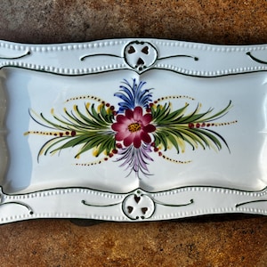 May include: Rectangular white ceramic serving tray with a hand-painted floral design. The tray features a central floral arrangement with a burgundy flower, green leaves, and accents of blue, yellow, and purple. The edges are scalloped and trimmed with green.