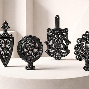 May include: Four black cast iron trivets in various shapes: a shield, a circle with a floral design, a paddle shape, and a tree. Each trivet has an intricate, openwork design. The trivets are displayed on a white surface.
