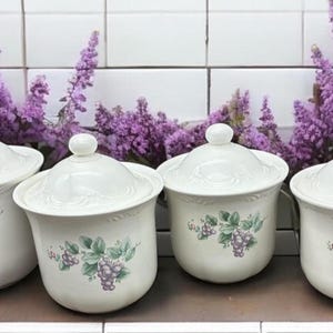 May include: Four white ceramic canisters with lids, each adorned with a grape cluster design in shades of green and purple. The canisters are arranged in a row, with a backdrop of purple flowers.