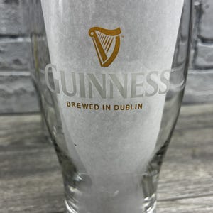 K&ouml;nnte beinhalten: Ein klares Pintglas mit dem Guinness-Logo und -Text. Das Logo ist eine goldene Harfe, und das Wort "GUINNESS" ist darunter in einer klaren Schrift gedruckt. Der Text "BREWED IN DUBLIN" ist unter dem Wort "GUINNESS" gedruckt.