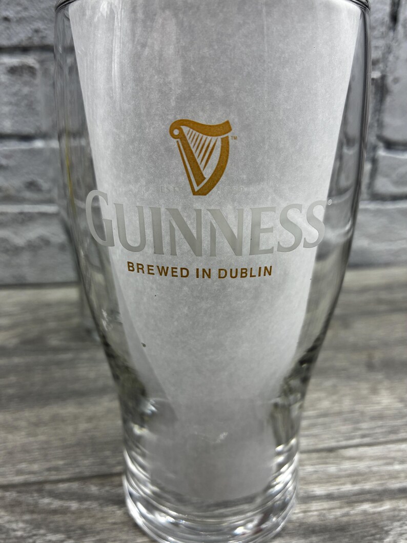 K&ouml;nnte beinhalten: Ein klares Pintglas mit dem Guinness-Logo und -Text. Das Logo ist eine goldene Harfe, und das Wort "GUINNESS" ist darunter in einer klaren Schrift gedruckt. Der Text "BREWED IN DUBLIN" ist unter dem Wort "GUINNESS" gedruckt.