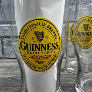 K&ouml;nnte beinhalten: Klare Biergl&auml;ser mit dem Guinness-Logo. Das Logo ist ein gelbes Oval mit schwarzem Text und einem Harfensymbol. Der Text lautet "Guinness Extra Stout" und "Traditionally Brewed".