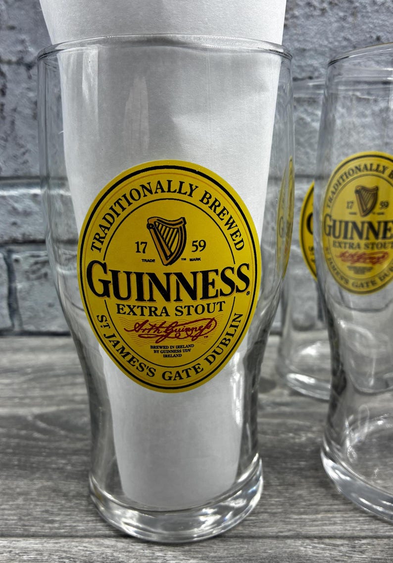 K&ouml;nnte beinhalten: Klare Biergl&auml;ser mit dem Guinness-Logo. Das Logo ist ein gelbes Oval mit schwarzem Text und einem Harfensymbol. Der Text lautet "Guinness Extra Stout" und "Traditionally Brewed".