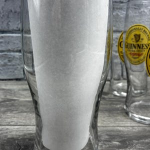 K&ouml;nnte beinhalten: Klare Biergl&auml;ser mit dem Guinness-Logo. Die Gl&auml;ser sind leer und haben einen Papiereinsatz. Der Hintergrund ist eine graue Holzoberfl&auml;che und eine graue Backsteinmauer.
