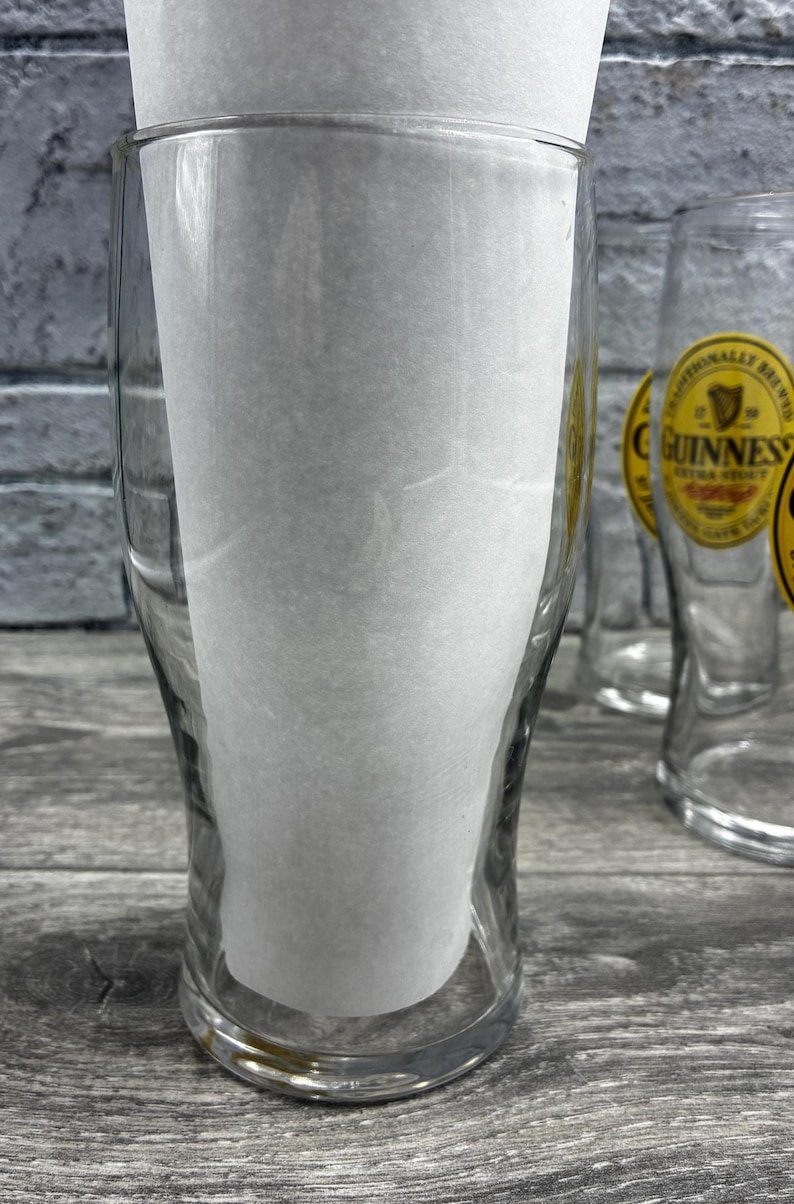 K&ouml;nnte beinhalten: Klare Biergl&auml;ser mit dem Guinness-Logo. Die Gl&auml;ser sind leer und haben einen Papiereinsatz. Der Hintergrund ist eine graue Holzoberfl&auml;che und eine graue Backsteinmauer.