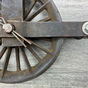 May include: Close-up of a weathered, antique metal pulley wheel. The wheel has multiple spokes and is attached to a rectangular metal frame. A cotter pin secures the wheel's axle. The metal has a rusty, aged appearance.
