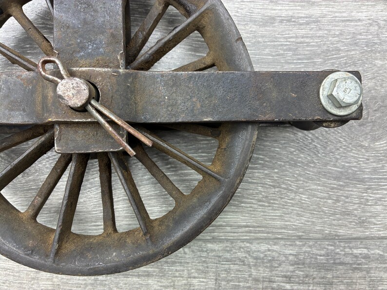 May include: Close-up of a weathered, antique metal pulley wheel. The wheel has multiple spokes and is attached to a rectangular metal frame. A cotter pin secures the wheel's axle. The metal has a rusty, aged appearance.