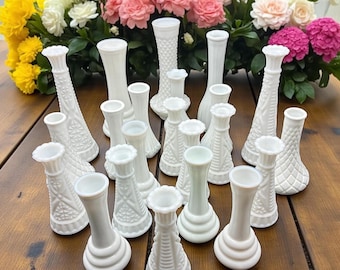 May include: A collection of white milk glass bud vases in various shapes and sizes, arranged on a wooden surface. The vases have textured and patterned designs. A colorful floral arrangement of yellow, pink, and white flowers is in the background.