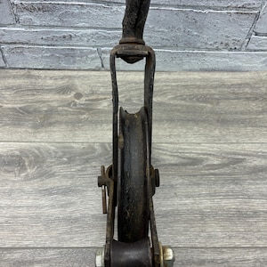 May include: An antique, black metal pulley system with a curved top and a wheel. The pulley has a U-shaped frame and is attached with bolts. The wheel is set in the center of the frame. The pulley is designed for lifting or pulling heavy objects.