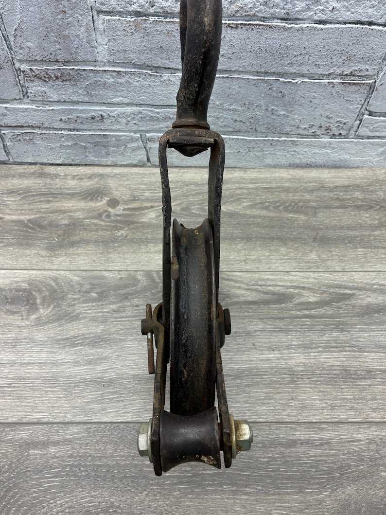 May include: An antique, black metal pulley system with a curved top and a wheel. The pulley has a U-shaped frame and is attached with bolts. The wheel is set in the center of the frame. The pulley is designed for lifting or pulling heavy objects.