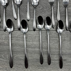 May include: An overhead shot of stainless steel silverware, including spoons and forks, arranged on a gray wooden surface. The spoons are in the foreground, with the forks and more spoons in the background. The silverware is reflective.