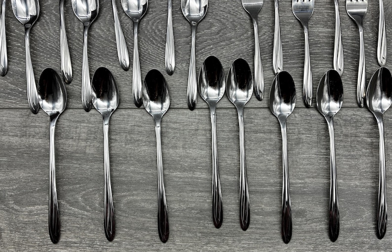 May include: An overhead shot of stainless steel silverware, including spoons and forks, arranged on a gray wooden surface. The spoons are in the foreground, with the forks and more spoons in the background. The silverware is reflective.