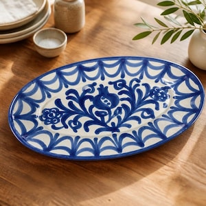 May include: An oval ceramic serving platter featuring a hand-painted blue floral and pomegranate motif centered on a cream background, surrounded by a decorative scalloped border pattern along the outer rim of the dish.