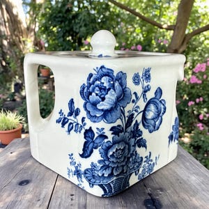 May include: A white ceramic square teapot with a lid and handle, adorned with a blue floral pattern. The design features large peony-like flowers and foliage. The teapot is sitting on a wooden surface.