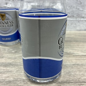May include: Clear glass with a blue and silver design. The glass has the Guinness Black Lager logo and text. The glass has a blue band at the bottom and a blue and white band at the top. The glass is cylindrical and appears to be a pint glass.