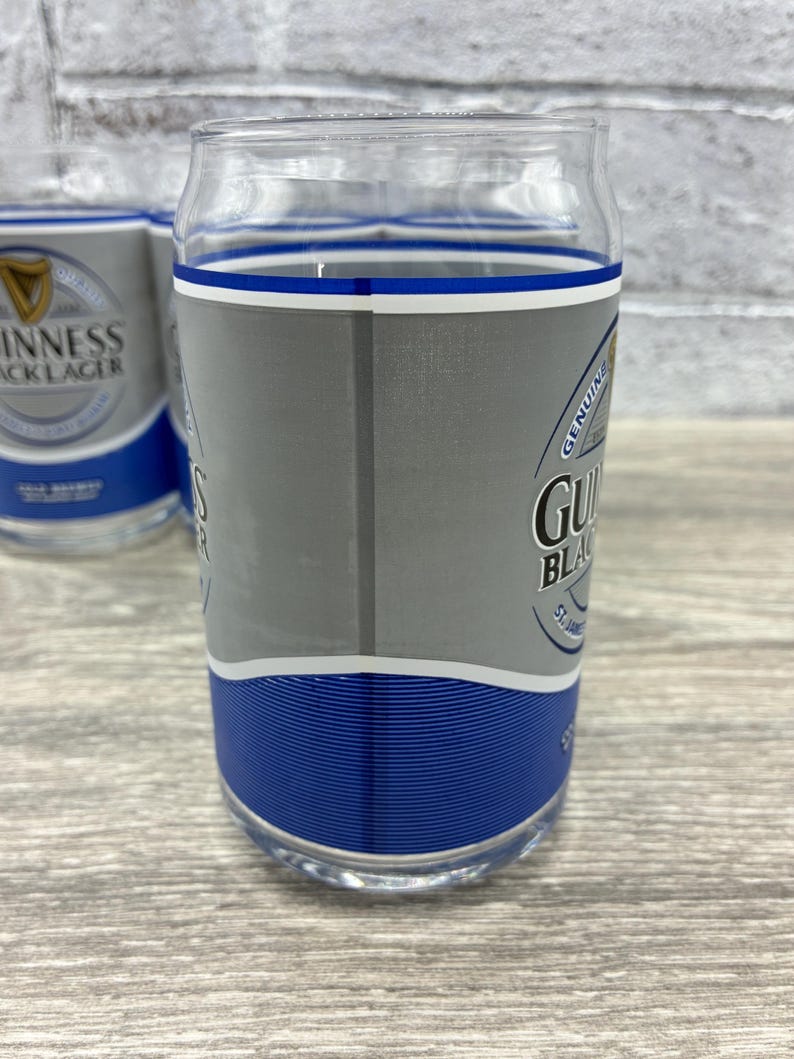 May include: Clear glass with a blue and silver design. The glass has the Guinness Black Lager logo and text. The glass has a blue band at the bottom and a blue and white band at the top. The glass is cylindrical and appears to be a pint glass.