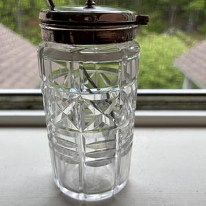 May include: A clear, cut-glass sugar bowl with a silver-colored lid and a small knob on top. The bowl has a geometric pattern and a small spoon inside. The sugar bowl is sitting on a white surface near a window.