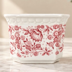 May include: A white ceramic planter with a scalloped rim and a repeating red floral pattern featuring small blossoms and leaves. The vessel has an octagonal shape with a wide opening and a tapered base, decorated with a delicate botanical print.