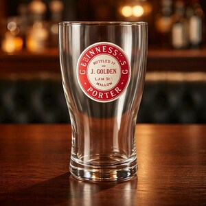 K&ouml;nnte beinhalten: Ein klares Pintglas mit einem rot-wei&szlig;en Guinness-Logo. Das Logo enth&auml;lt die Worte "Guinness", "Bottled by J. Golden" und "Porter". Das leere Glas steht auf einer Holzoberfl&auml;che.