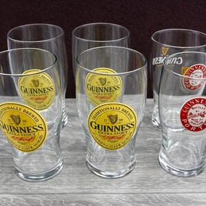 K&ouml;nnte beinhalten: Sechs klare Pintgl&auml;ser mit dem Guinness-Logo. Die Gl&auml;ser haben eine gebogene Form und einen breiten Boden. Das Guinness-Logo ist ein gold-schwarzes Etikett mit den Worten "Guinness Extra Stout" und einem Harfensymbol.