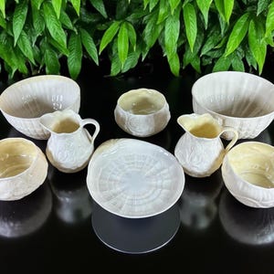 May include: A collection of cream-colored ceramic serving pieces, including bowls, pitchers, and a plate. The pieces have a textured, possibly embossed, design. The arrangement suggests a dining or serving set.