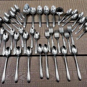May include: An assortment of stainless steel silverware, including spoons, forks, and a ladle, arranged on a wooden surface. The silverware has a shiny, reflective surface, and the arrangement suggests a formal dining setting.