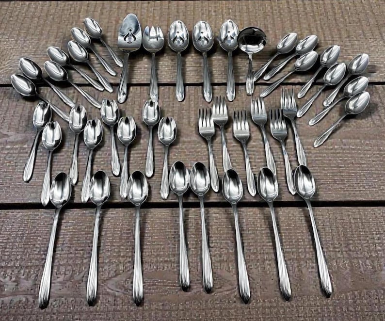 May include: An assortment of stainless steel silverware, including spoons, forks, and a ladle, arranged on a wooden surface. The silverware has a shiny, reflective surface, and the arrangement suggests a formal dining setting.