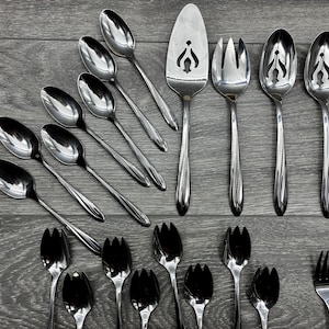 May include: A collection of stainless steel silverware, including spoons, forks, and serving utensils, arranged on a gray wooden surface. The spoons have a classic design, and the serving pieces have decorative cutouts.