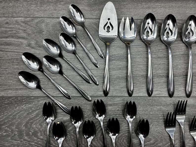 May include: A collection of stainless steel silverware, including spoons, forks, and serving utensils, arranged on a gray wooden surface. The spoons have a classic design, and the serving pieces have decorative cutouts.