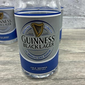 May include: Clear glass pint glasses with a gray and blue design. The glasses feature the Guinness Black Lager logo, a harp, and the text "Genuine Quality" and "Cold Brewed with Roasted Barley."