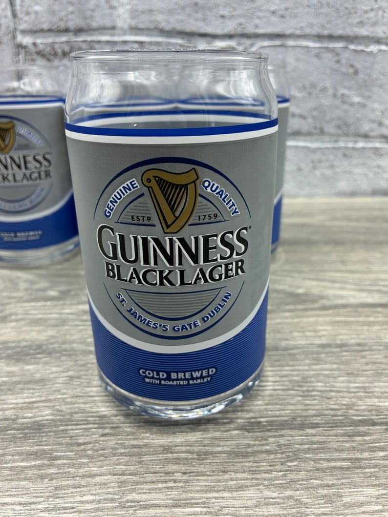 May include: Clear glass pint glasses with a gray and blue design. The glasses feature the Guinness Black Lager logo, a harp, and the text "Genuine Quality" and "Cold Brewed with Roasted Barley."