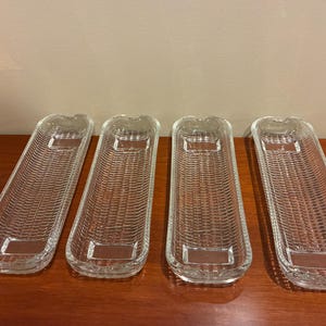 May include: Four clear glass serving dishes with a woven basket texture. Each dish is rectangular with a slightly curved shape and a small handle at one end. The dishes are arranged on a wooden surface.