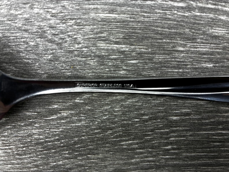 May include: Close-up of a stainless steel fork with the text "SUPERIOR STAINLESS USA" etched on the handle. The fork is set against a textured, gray wooden surface.