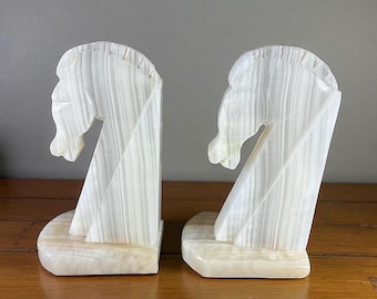 May include: A pair of white onyx bookends, each sculpted in the shape of a horse's head. The bookends have a vertical design with a base and are set against a neutral background.